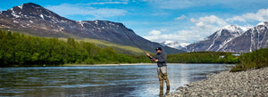 Man spey casting on a river with mountains in the background with a partly cloudy blue sky.